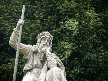 Statue d'un druide barbu tenant un bâton, devant un feuillage