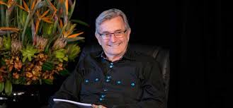 Homme souriant aux lunettes, assis devant un bouquet de fleurs, vraisemblablement Gary Douglas